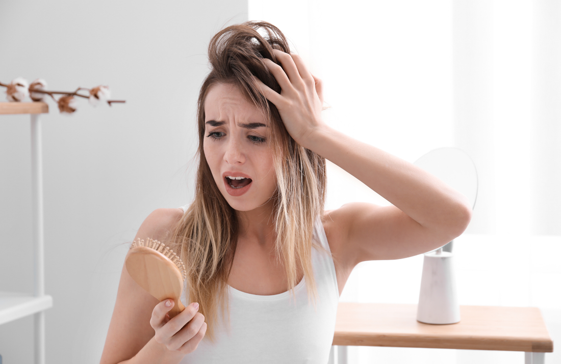 young woman worried about hair loss holding a hairbrush with strands of hair in a bright room, illustrating natural hair oils for growth benefits