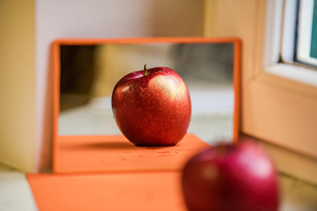 Red apple reflected in a small orange mirror on a table by the window, symbolizing the perks of apple cider vinegar for health and wellness.
